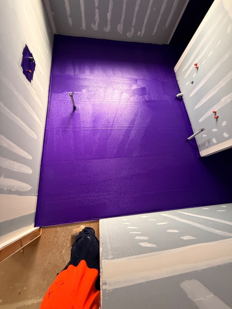 Purple waterproofing membrane fully applied to bathroom floor with plumbing rough-in visible and tradesman in hi-vis on site — Keysborough job by Casey Quality Coatings