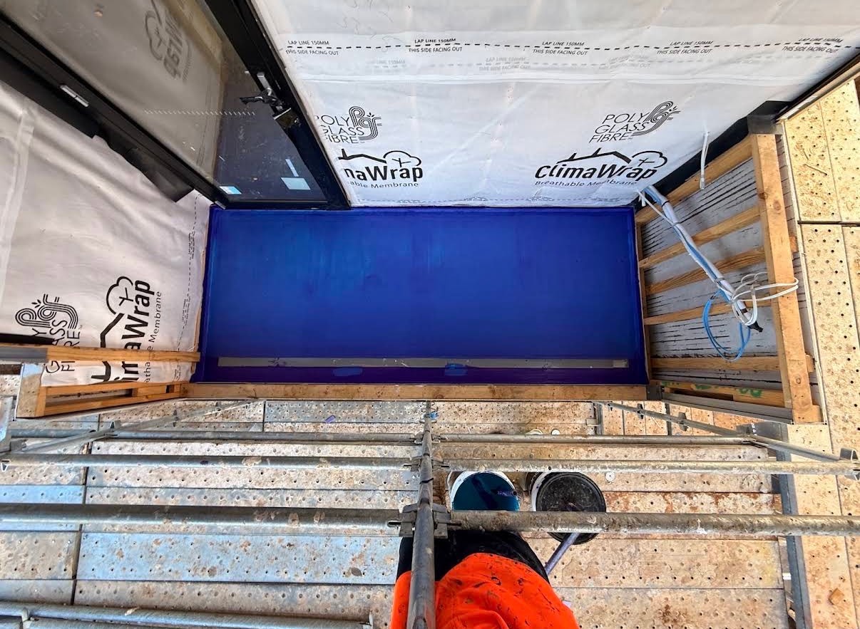 Overhead view from scaffolding of balcony deck with vivid blue waterproofing membrane fully applied, ClimaWrap breathable membrane on timber framing surround with 150mm lap lines marked, tradesman in hi-vis orange visible — Noble Park new-build by Casey Quality Coatings